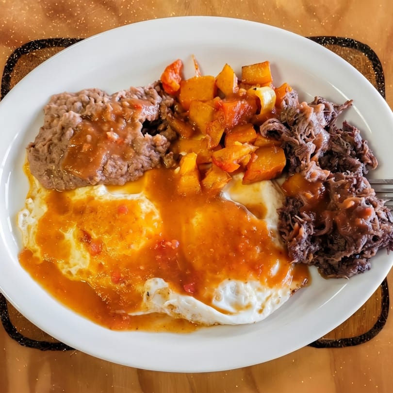 A white plate with two fried eggs, refried beans, shredded beef, and diced potatoes, all covered in red sauce.