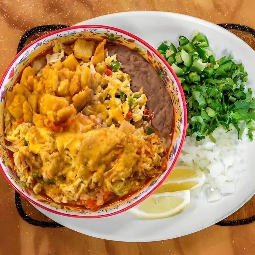 A colorful bowl of cheesy chilaquiles with refried beans, alongside chopped cilantro, onions, and lemon wedges.