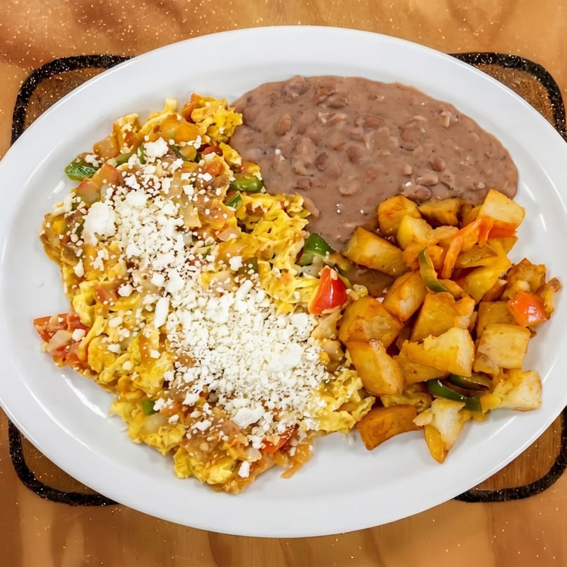 A white plate holds scrambled eggs with cheese, refried beans, and seasoned potato chunks.