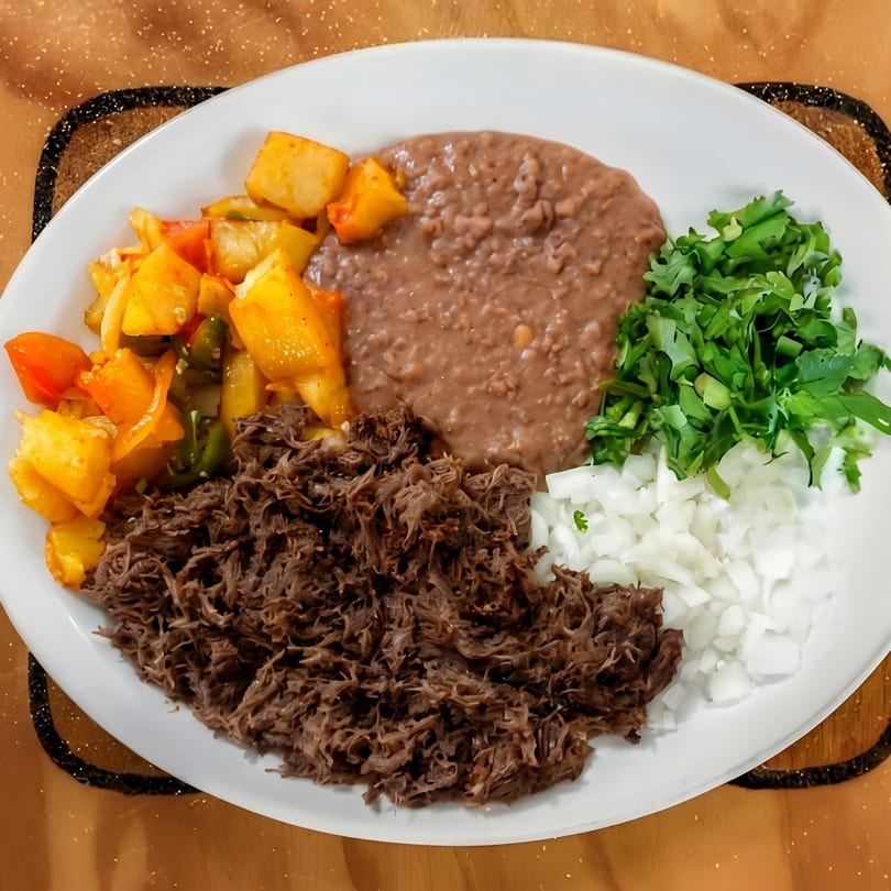 A white plate with shredded beef, refried beans, diced potatoes, cilantro, and chopped onions.